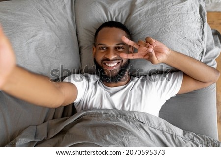 Overhead above top view portrait of relaxed black man lying in bed at home after waking up, taking selfie showing peace sign v victory gesture with 2 fingers, holding cellphone looking at camera