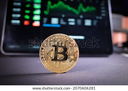 a bitcoin coin is standing on a dark background and in the background is the bitcoin chart