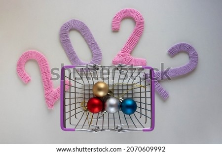 The numbers 2022, made in pink and lilac threads on a white background, are next to a grocery basket with Christmas tree toys. The concept of the New Year.