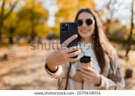 attractive young woman walking in autumn park taking pictures using smartphone, wearing checkered coat, sunglasses, happy mood, fashion style trend