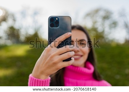 portrait of candid attractive young smiling woman in pink sweater walking in green park holding phone taking pictures happy mood, fashion style trend