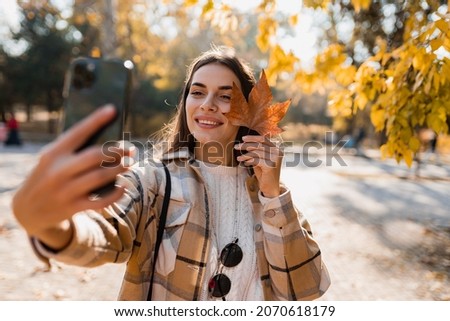 attractive young smiling woman walking in autumn park taking selfie pictures with leaf using smartphone, wearing checkered coat, happy mood, fashion style trend