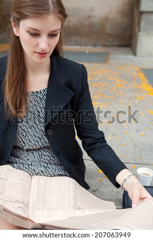 Close up portrait view of an attractive professional young woman sitting on stone steps in the city reading a financial newspaper, outdoors. Business communications and information.