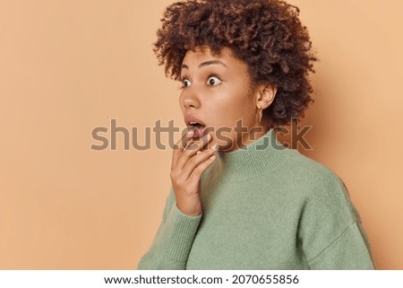 Startled curly haired woman holds chin and stares bugged eyes away holds breath from amazement notices something terrible wears casual jumper isolated over beige background blank copy space. Royalty-Free Stock Photo #2070655856