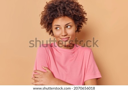 Horizontal shot of pleased young woman with curly hair looks away being deep in thoughts wears casual pink t shirt isolated over beige background recalls nice memory. People and thoughts concept