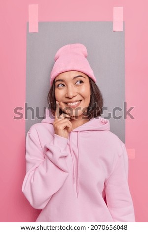 Dreamy Asian woman keeps finger on cheek smiles plesantly thinks about something pleasant has smile on face dressed in hoodie and hat pictures something in mind poses against blank copy space