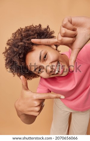 Young positive woman makes camera frame with fingers tilts head and smiles happily searches perfect angle has playful mood poses indoor against brown background uses imagination pictures something