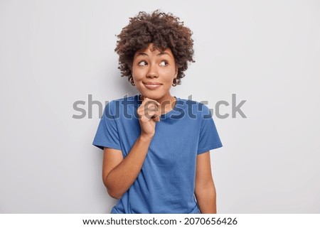 Horizontal shot of dreamy young curly haired woman looks away with smiling face keeps hand under chin wears casual blue t shirt pictures something beautiful in mind poses against white background