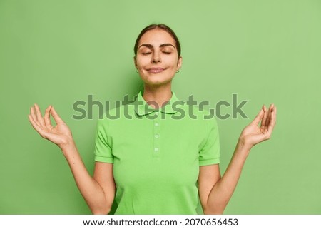 Relaxed beautiful woman keeps fingers together for meditation makes zen mudra sign breathes patient inhales positive vibes closes eyes closed wears casual t shirt isolated over green background