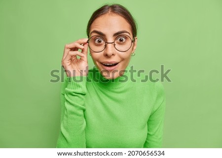 Photo of surprised cheerful woman with dark hair looks wondered through spectacles cannot believe her eyes dressed in casual jumper isolated over vivid green background. Human reactions concept Royalty-Free Stock Photo #2070656534