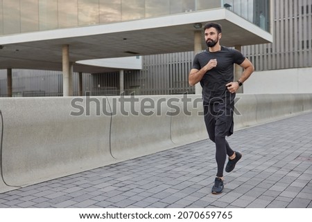 Determined bearded adult man in activewear jogs in city street covers long distance enjoys morning running exercises outdoors keeps fit and healthy muscular strength flexibility physical power Royalty-Free Stock Photo #2070659765
