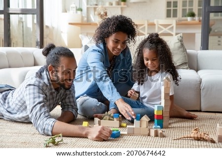 Happy millennial Black couple of parents and cute daughter kid playing on heating carpeted floor at cozy home, constructing town, buildings, towers from toy wooden bricks. Family game activity Royalty-Free Stock Photo #2070674642