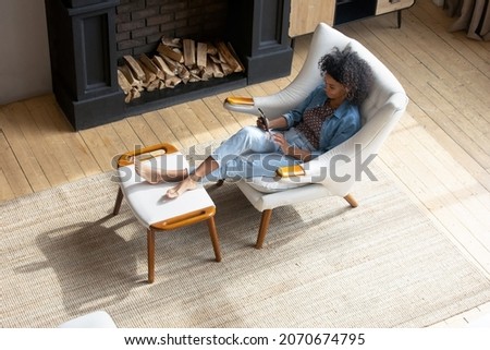 Relaxed young African American woman resting in comfortable armchair with legs on footrest chair, relaxing at cozy home fireplace, holding mobile phone, using online app on mobile phone. High angle Royalty-Free Stock Photo #2070674795