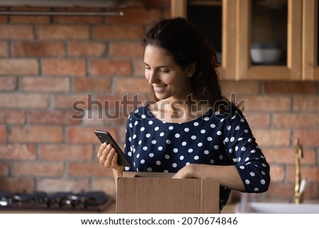 Young hispanic woman blogger unpack postal delivery container using mobile phone camera to film unboxing process shoot pictures. Happy latina female get gift by mail call friend to thank for present