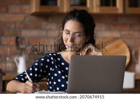 Smiling millennial latin woman tutor teacher wear glasses use laptop to search new material for lesson online write notes in daybook. Confident young female student take test exam at elearning course