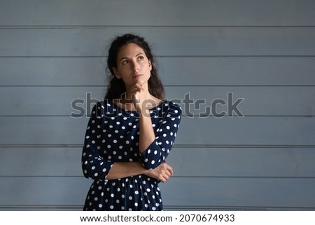 Thoughtful young latin woman stand close to grey wall look up ponder on interesting offer proposal imagine result. Puzzled millennial lady touch chin reflect on idea make choice decision. Copy space