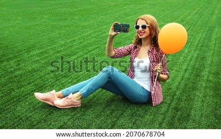 Happy smiling young woman taking a selfie by smartphone with balloon sitting on the grass in a summer park