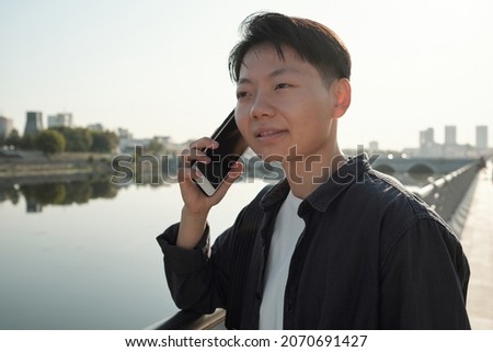 Pretty Chinese business agent with smartphone by her ear consulting one of clients while standing by riverside