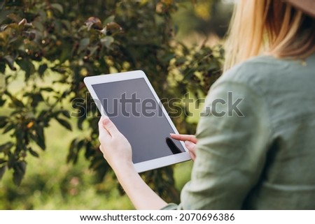 A farmer with a tablet in her hands stands in green garden. Woman smiling, looking at tablet screen. Modern lifestyle and relaxation concepts. Mock up