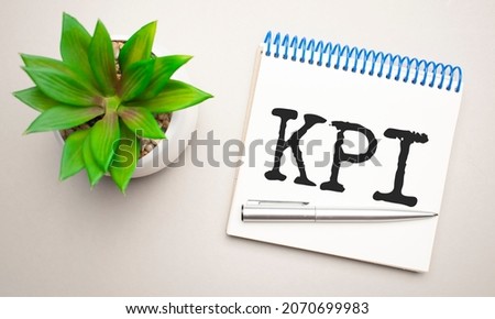 Business kpi is written in a white notepad near a clipboard, calculator, green plant, glasses and a pen on a yellow and concrete background. Business concept. Flat lay.