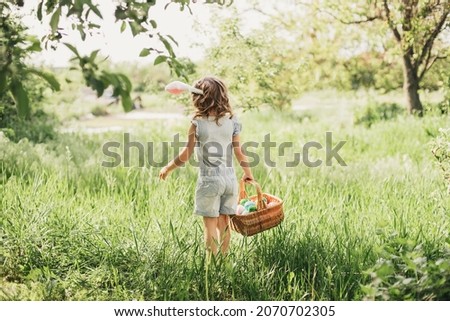 Easter egg hunt. Girl child Wearing Bunny Ears Running To Pick Up Egg In Garden. Easter tradition. Baby with basket full of colorful eggs. Back view