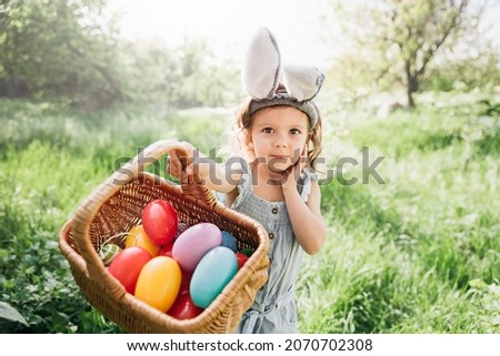 Easter egg hunt. Girl child Wearing Bunny Ears Running To Pick Up Egg In Garden. Easter tradition. Baby with basket full of colorful eggs. wide angle view