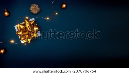 Christmas composition. White gift box with golden ribbon, New Year balls and sparkling lights garland in xmas composition on black background for greeting card. Flat lay, top view, copy space