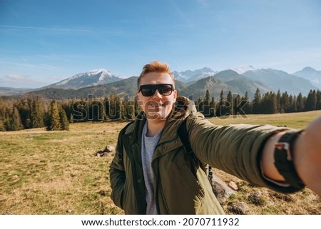 Young man in sunglasses making selfie photo high in the snow mountains enjoying the view. Freedom, happiness, travel and vacations concept, outdoor activities, he wearing a green jacket