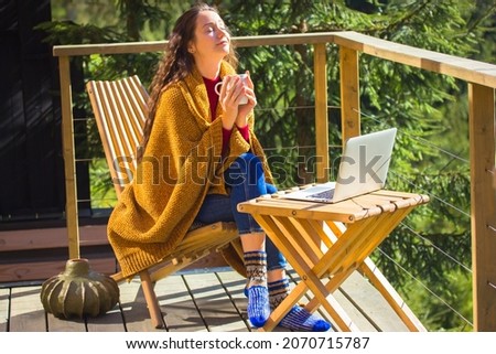 Pretty white girl is sitting relaxing on balcony wrapped in yellow plaid holding cup of tea, enjoying coffee. Young brunette woman works online, remotely on laptop. Sweet home in a wood at autumn day. Royalty-Free Stock Photo #2070715787