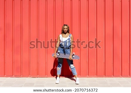 Black woman dressed casual, wtih a skateboard on red urban wall background.