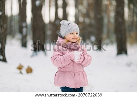 Close up portrait of a little cute girl in pink clothes with a cup of hot tea, posing outdoors on the background of beautiful winter snowy forest, looking away