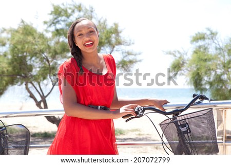Colorful portrait of a young and attractive african american teenager girl with her bicycle, joyfully relaxing by the sea during a sunny day on holiday. Active and healthy lifestyle, outdoors.