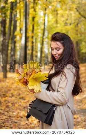 Brunette woman in autumn park with a cup of coffee and autumn leaves. Small black bag. Autumn mood. Yellow, red and green leaves.