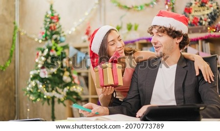 Young business team are celebrating holiday in workplace are sitting in Santa hats and Give a gift to the colleagues.Beautiful young couple in love at office, celebrating with a gift box exchange.