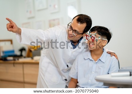 Ophthalmologist or optical staff point to the direction for viewing of Indian boy who wear special glasses to test his eye during process of eyecare in the shop. Royalty-Free Stock Photo #2070775040