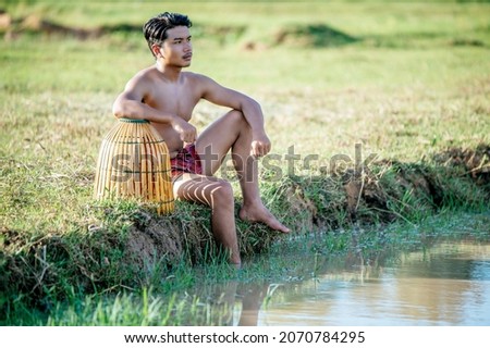 Portrait Young man topless use bamboo fishing trap to catch fish for cooking, Asian young farmer man in rural lifestyle, blurred buffalo is soaking in the swamp. in background