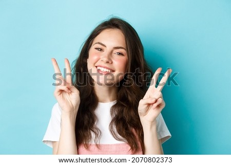 Close up portrait of attractive positive girl saying cheeze, smiling and showing v-signs peace, standing over blue background