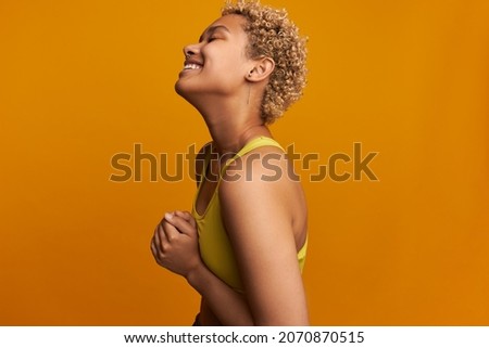 Profile picture of adorable, charming, stylish African female with blonde short curly hair wearing yellow fitness top, laughing putting head back pressing fists against belly, having fun doing workout