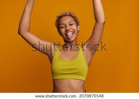 Funny cropped picture of cheerful carefree young blonde woman of mixed race holding hands high, sticking tongue, fooling around making faces, having cute short poodles, wearing yellow stylish top