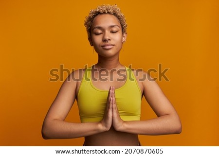 Meditation, relaxation, reaching zen, spiritual practice. Charming young blonde Afro female standing in namaste position, keeping eyes closed, breathing deeply, feeling harmony inside and outside
