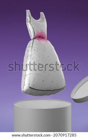 Shot of a balloon shaped as a pretty gray dress with pink flowers. The silver balloon is flying out of a gray box. The photo is taken on a violet background.