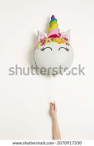 Shot of a balloon shaped as a unicorn head. It has a rainbow horn and multicolored patterns on the top of the head. A white balloon is held by a hand of a person on a white background.