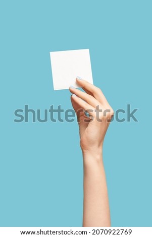 Female hand with blue manicure holding a white blank message card. Paper note in female model hand on blue background. Copy space.
