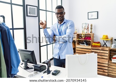 Young african man working as manager at retail boutique with a big smile on face, pointing with hand finger to the side looking at the camera. 