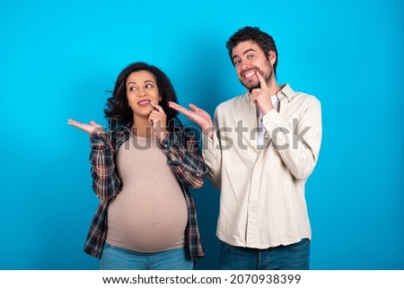 Positive young couple expecting a baby standing against blue background advert promo touch finger teeth