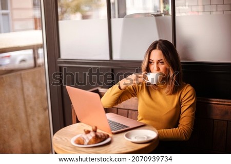 Young woman freelancer drinking coffee and eating croissant while working remotely on laptop computer in cafe, caucasian female enjoying breakfast during online remote work in coffeeshop Royalty-Free Stock Photo #2070939734