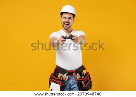 Young fun gambling employee handyman man in protective helmet hardhat play pc game with joystick console isolated on yellow background Instruments accessories renovation apartment room Repair concept