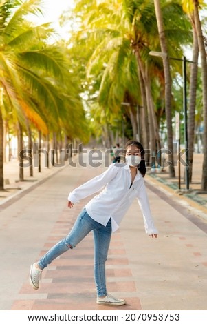 A young Asian woman wearing a white shirt and jeans is posing for a photo on the Bangsaen beach walkway on a day when there are not many people behind the coconut trees. On a clear day, Thailand