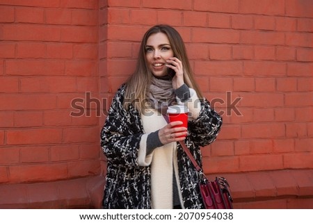 Young smiling woman with phone and coffee on a city street. Beautiful blonde in a warm stylish coat against a background of a brick wall. Blogging and remote work.