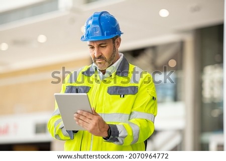 Serious and focused architect or engineer working on construction site using digital tablet. Mature man working on digital tablet at construction site. Mid adult manual worker checking list on laptop. Royalty-Free Stock Photo #2070964772